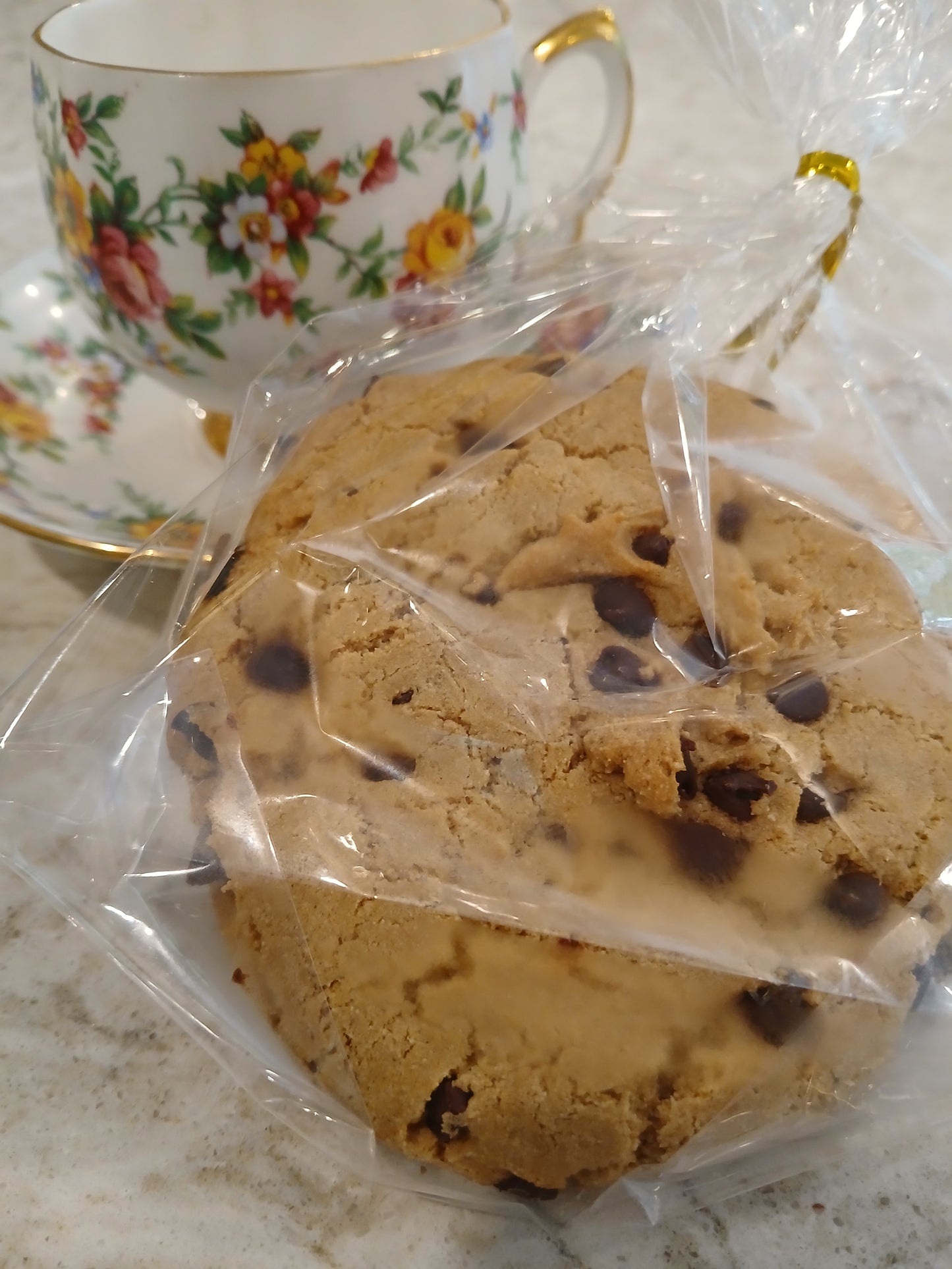 Chocolate chip cookies in clear plastic wrap with a floral teacup in the background.
