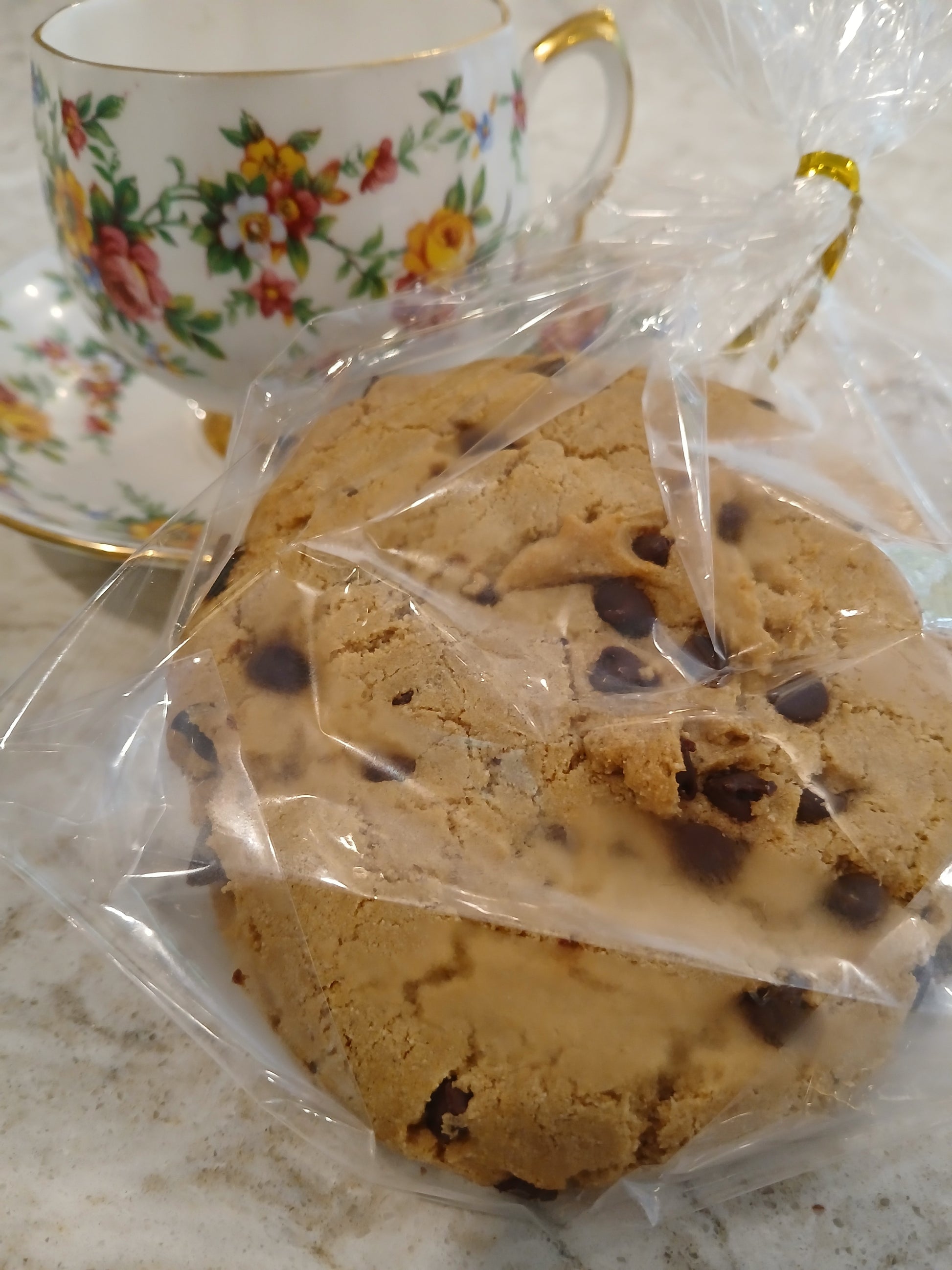 Chocolate chip cookies in clear plastic wrap with a floral teacup in the background.
