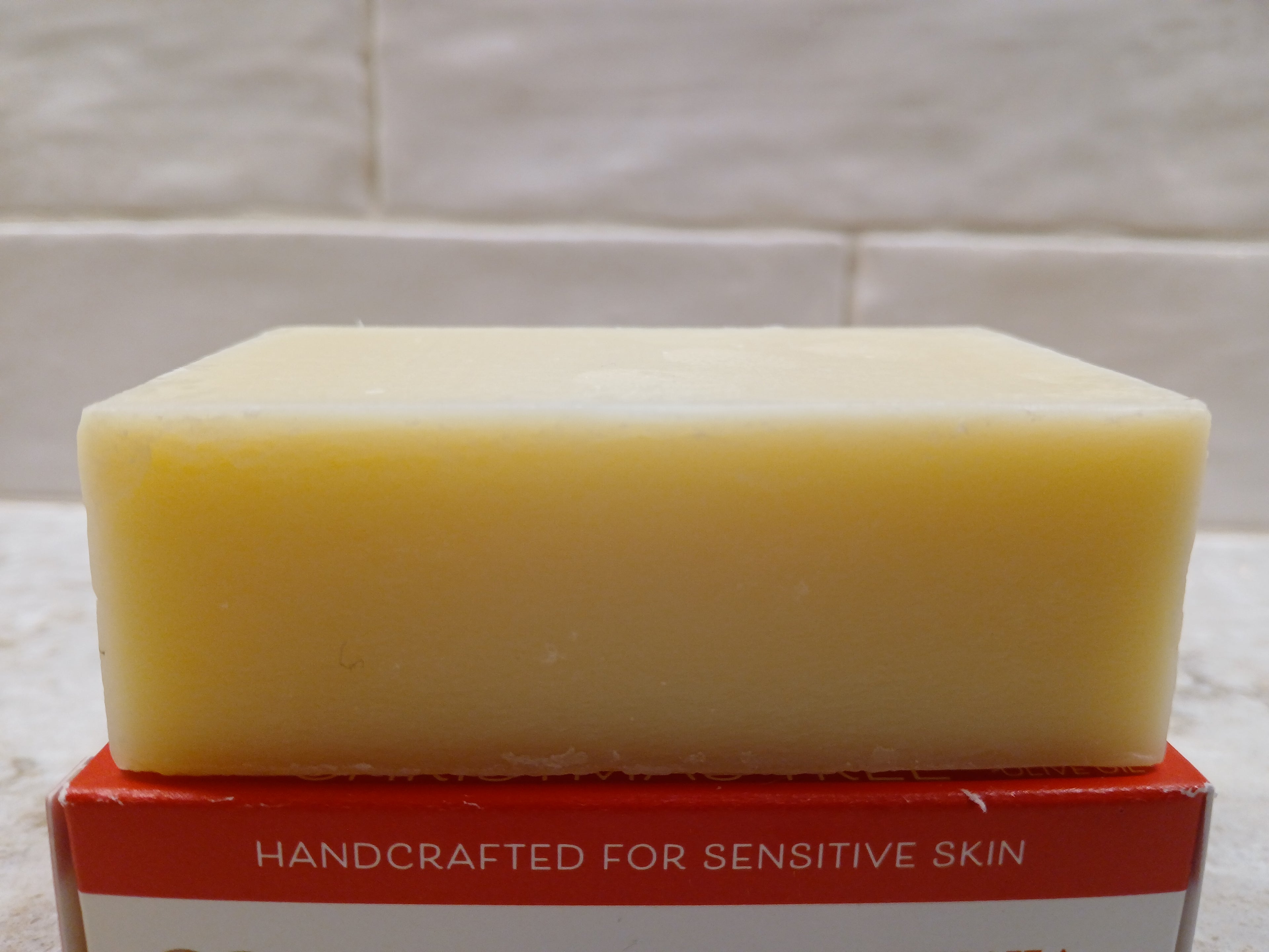 Bar of lemongrass soap on a red box with 'Handcrafted for Sensitive Skin' text, against a tiled bathroom wall background.