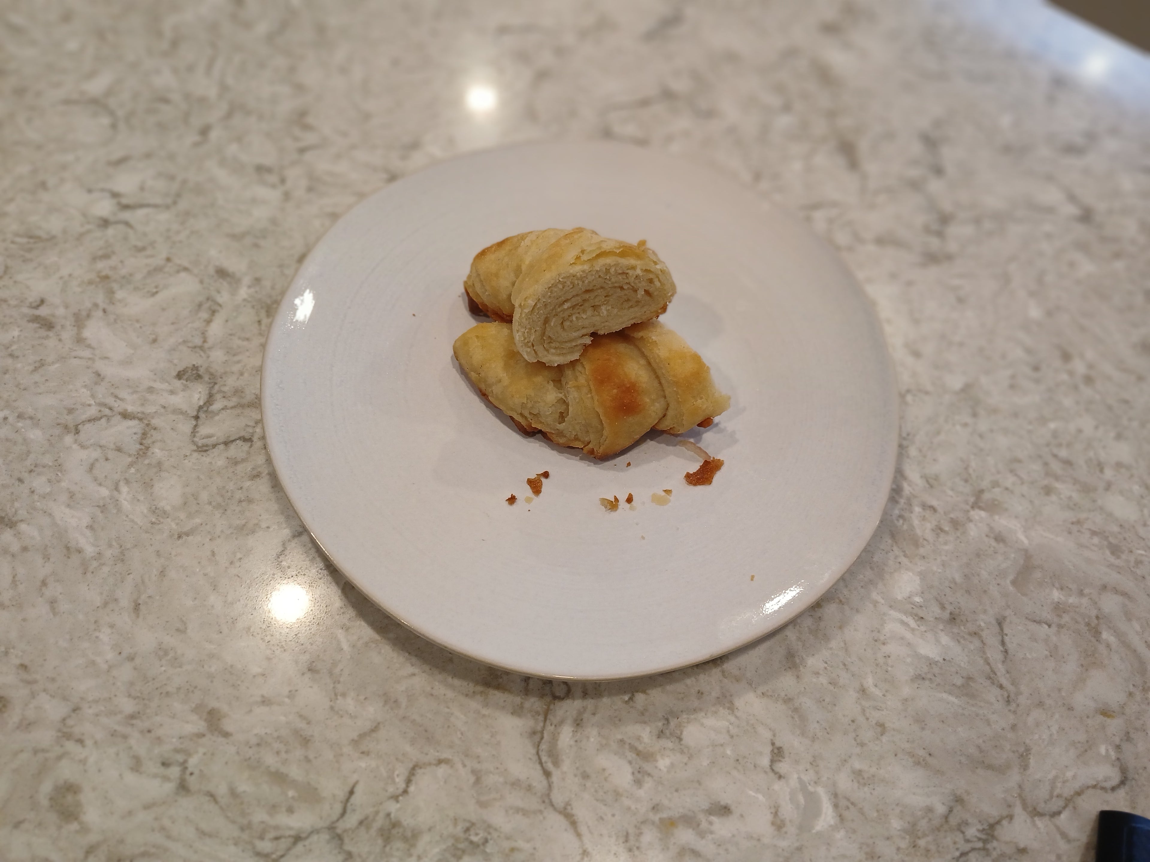 Two gluten-free croissants on a white plate with crumbs on a marble countertop