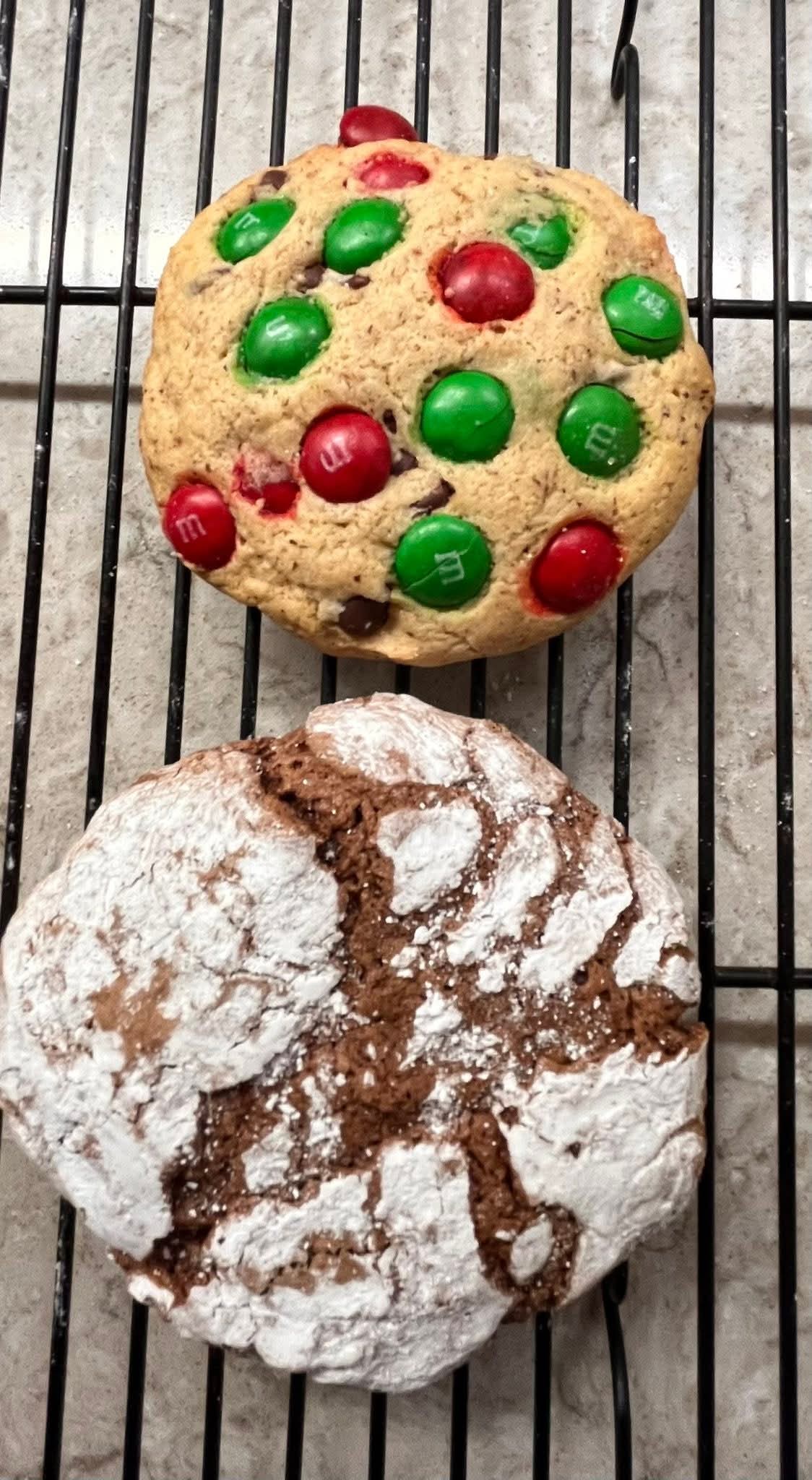 Two gluten-free cookies on a cooling rack, one chocolate crinkle cookie with powdered sugar and the other M&M cookie.